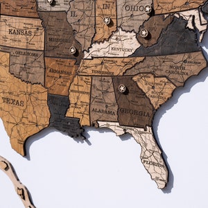 May include: A wooden map of the United States with state names and pin markings. The map is made of different shades of brown wood and has a light brown background.