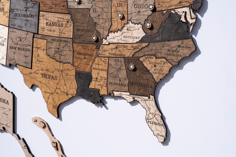 May include: A wooden map of the United States with state names and pin markings. The map is made of different shades of brown wood and has a light brown background.