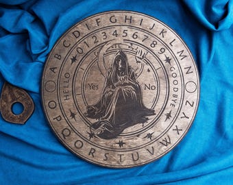 Może przedstawiać: Drewniana plansza Ouija ze szczegółową ilustracją postaci w kapturze, księżyca, gwiazd i alfabetu. Plansza zawiera cyfry 0-9 oraz słowa "Cześć", "Tak", "Nie" i "Do widzenia". W zestawie znajduje się również planchette.
