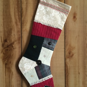 May include: A handmade Christmas stocking with a patchwork design. The stocking features various fabrics, including floral lace, red stripes, and plaid patterns. Decorative buttons and lace trim add detail. The stocking hangs against a wooden background.
