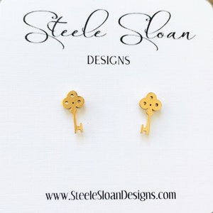 May include: A pair of gold key-shaped stud earrings on a white card with the text "Steele Sloan Designs" and "www.SteeleSloanDesigns.com".