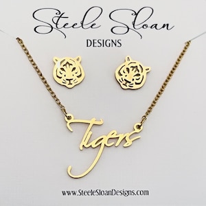 May include: Gold-colored jewelry set featuring a necklace with the word "Tigers" in cursive script and matching tiger head stud earrings. The brand name "Steele Sloan Designs" is visible at the top, with the website address at the bottom. The jewelry is displayed on a white background.