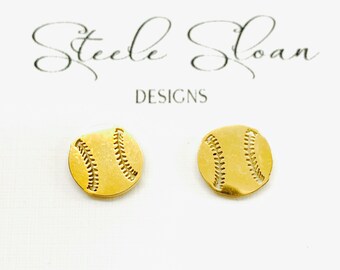 May include: A pair of gold-colored baseball stud earrings. Each earring is a small, circular design with stitched detailing, resembling a baseball. The earrings are displayed on a white card with the text "Steele Sloan Designs" and the website address.