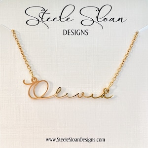 May include: Gold-colored necklace spelling the name "Olivia" in a cursive font. The necklace hangs on a white card with the text "Steele Sloan Designs" and the website address www.SteeleSloanDesigns.com.