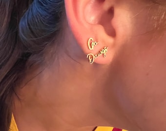 May include: A pair of gold earrings with the words "Go Dawgs" written on them. The earrings are worn in the left ear of a person wearing a maroon and gold shirt.