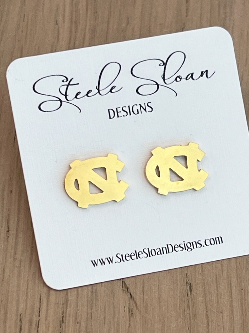 Op de afbeelding: Gouden oorbellen met het UNC Tar Heels logo. De oorbellen worden getoond op een witte kaart met de tekst "Steele Sloan Designs" en "www.SteeleSloanDesigns.com".