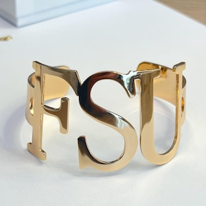May include: Gold-toned metal bracelet with the letters FSU cut out. The bracelet is open and ready to be worn.