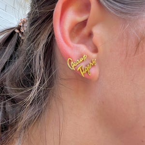 May include: Gold-colored stud earrings spelling out "Geaux Tigers" in a cursive font. The earrings are worn on an ear, with the text positioned to be read from left to right.