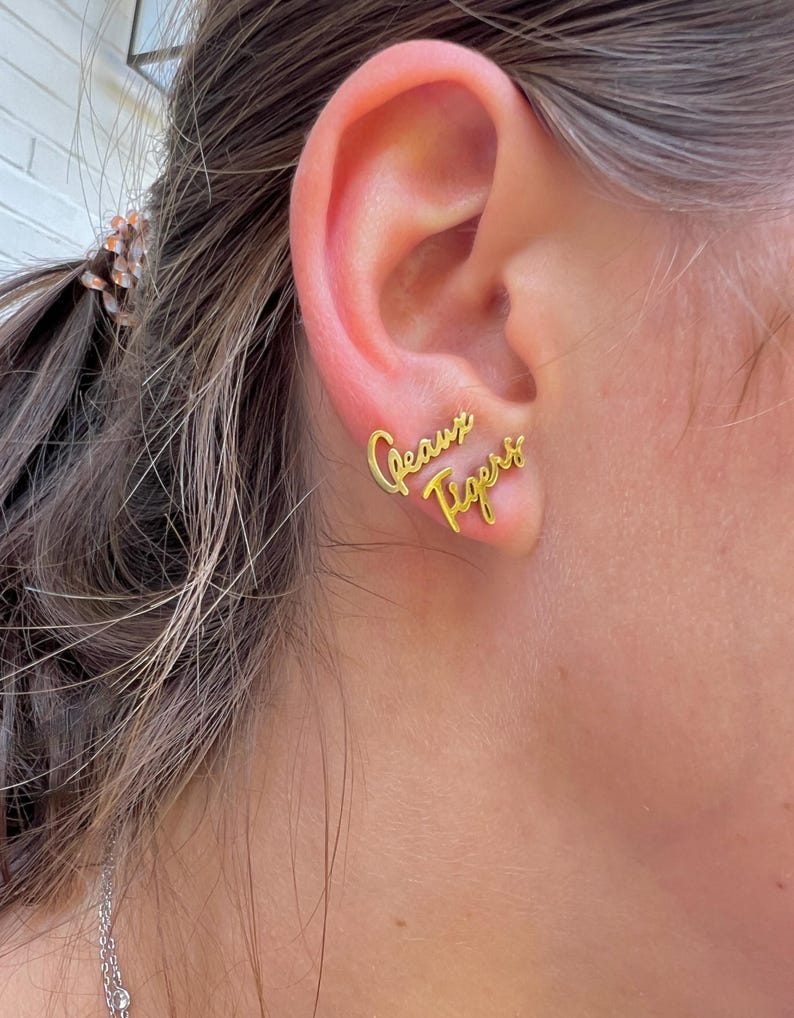 May include: Gold-colored stud earrings spelling out "Geaux Tigers" in a cursive font. The earrings are worn on an ear, with the text positioned to be read from left to right.