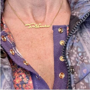 May include: A gold necklace with a cursive nameplate pendant that reads "Tricia". The necklace is worn by a person wearing a purple and floral patterned shirt.