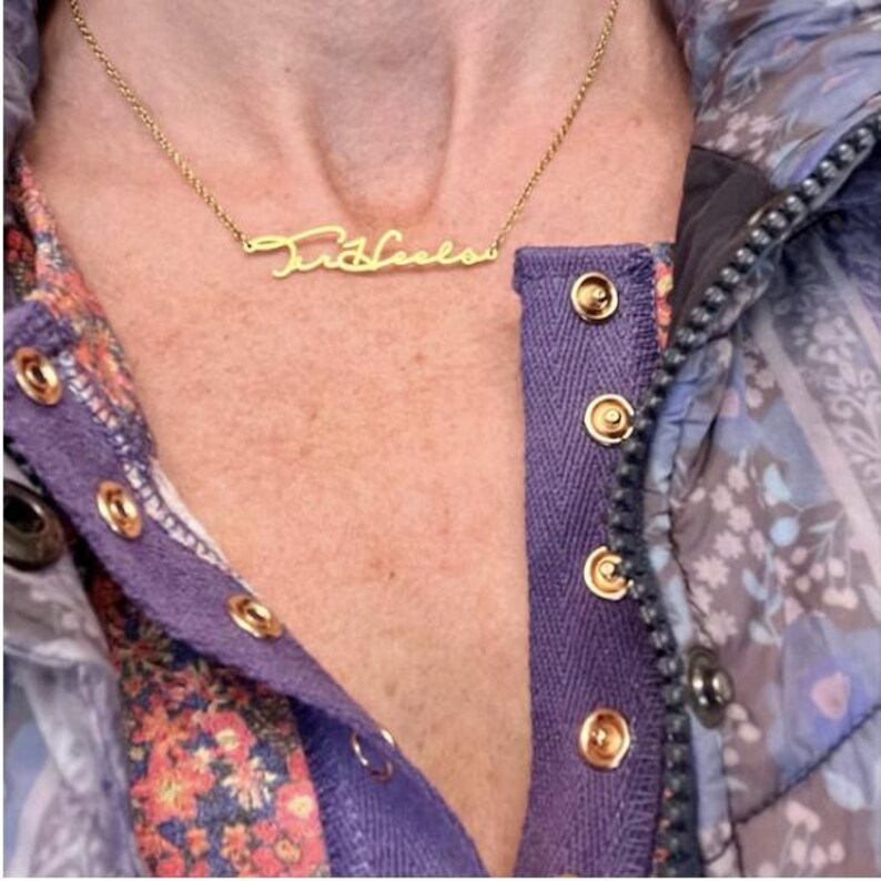 May include: A gold necklace with a cursive nameplate pendant that reads "Tricia". The necklace is worn by a person wearing a purple and floral patterned shirt.
