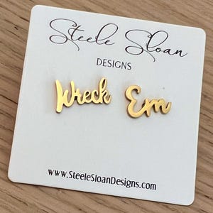 May include: Gold-colored "Wreck Em" stud earrings on a white card with the text "Steele Sloan DESIGNS" and the website address www.SteeleSloanDesigns.com. The earrings are in a cursive font.