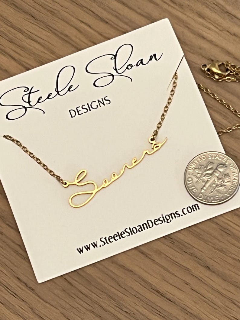 May include: A gold necklace with the word "Sooner" written in cursive script. The necklace is on a white card with the text "Steele Sloan Designs" and the website address "www.SteeleSloanDesigns.com".