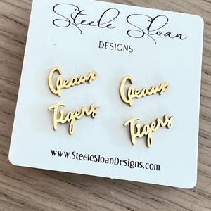 May include: Gold-colored stud earrings with the words "Geaux" and "Tigers" in a cursive font. The earrings are displayed on a white card with the text "Steele Sloan Designs" and a website address.
