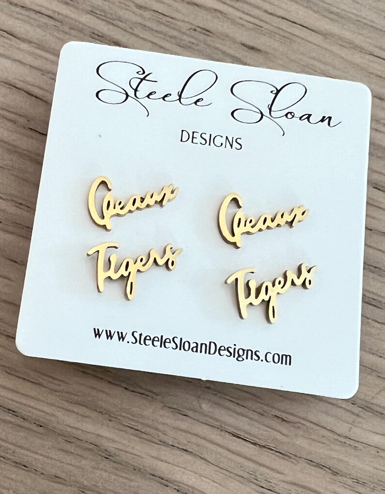 May include: Gold-colored stud earrings with the words "Geaux" and "Tigers" in a cursive font. The earrings are displayed on a white card with the text "Steele Sloan Designs" and a website address.