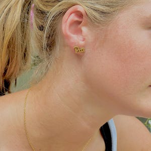 May include: Gold-colored stud earrings spelling out the word "Paris" in a cursive font. A delicate gold necklace with the letters "D", "I", "Y", and "T" is also visible. The jewelry is worn by a person with blonde hair.