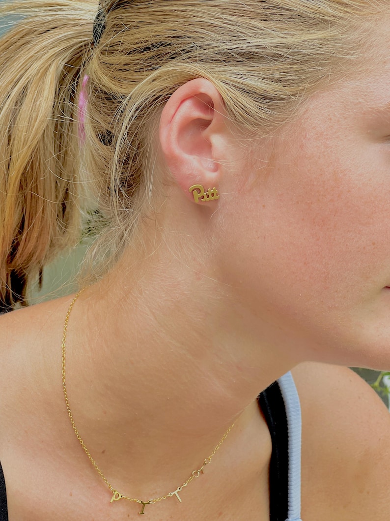 May include: Gold-colored stud earrings spelling out the word "Paris" in a cursive font. A delicate gold necklace with the letters "D", "I", "Y", and "T" is also visible. The jewelry is worn by a person with blonde hair.
