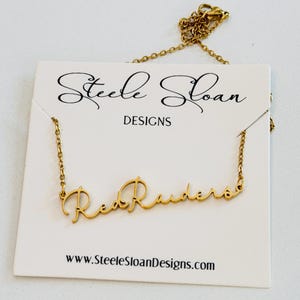May include: Gold-colored necklace with the word "Red Raiders" in a cursive font. The necklace is displayed on a white card with the text "Steele Sloan DESIGNS" and the website address.