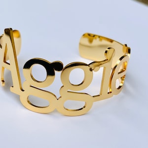 May include: A gold-colored cuff bracelet with the name "Aggie" cut out in a bold, stylized font. The bracelet has a smooth, polished finish and a simple, open design.