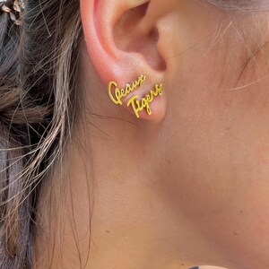 May include: Gold-colored stud earrings spelling out "Geaux Tigers" in a cursive font. The earrings are worn on an ear, with the text positioned to be read from left to right. The earrings are a stylish accessory.