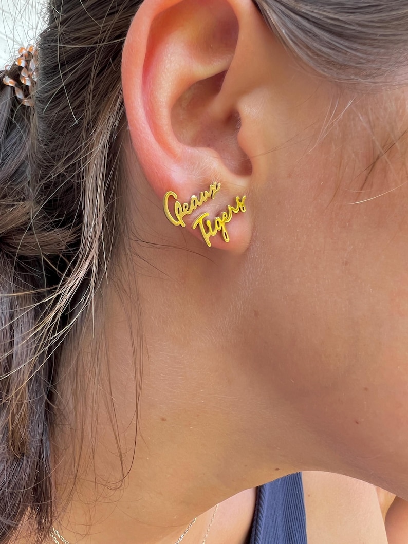 May include: Gold-colored stud earrings spelling out "Geaux Tigers" in a cursive font. The earrings are worn on an ear, with the text positioned to be read from left to right. The earrings are a stylish accessory.