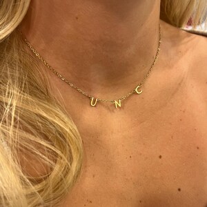 May include: A gold chain necklace with the letters "UNC" hanging from it. The necklace is worn by a person with blonde hair.