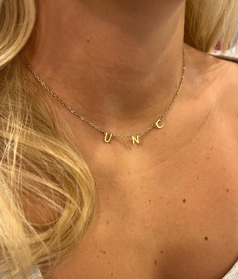 May include: A gold chain necklace with the letters "UNC" hanging from it. The necklace is worn by a person with blonde hair.