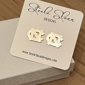 Op de afbeelding: Een paar zilveren oorbellen met het UNC Tar Heels logo. De oorbellen worden getoond op een witte kaart met de tekst "Steele Sloan Designs" en "www.SteeleSloanDesigns.com".
