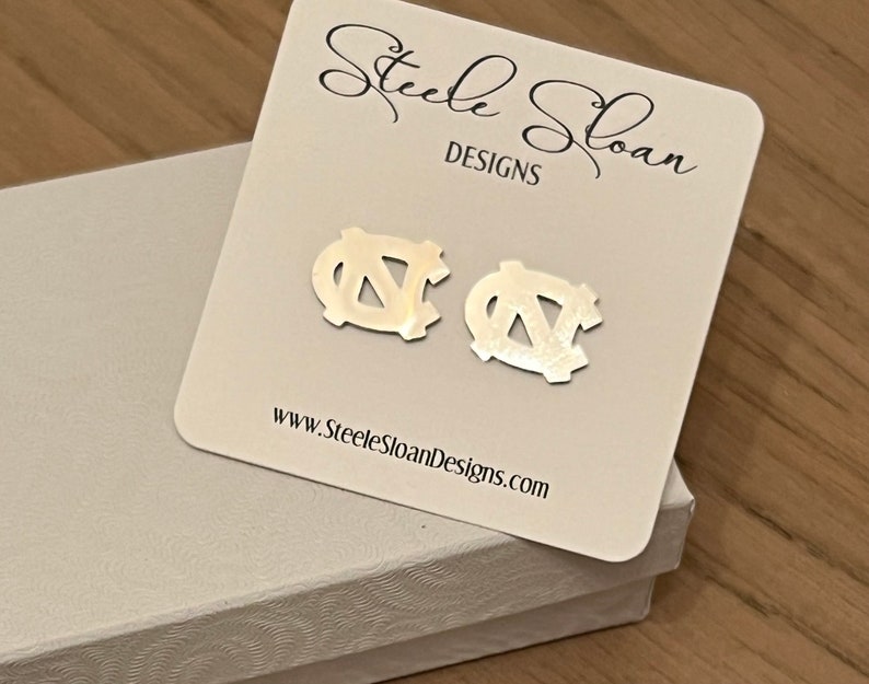 Op de afbeelding: Een paar zilveren oorbellen met het UNC Tar Heels logo. De oorbellen worden getoond op een witte kaart met de tekst "Steele Sloan Designs" en "www.SteeleSloanDesigns.com".