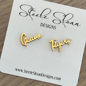 May include: Gold-colored "Geaux Tigers" earrings on a white card. The earrings are in a cursive font. The card has the text "Steele Sloan DESIGNS" and the website address www.SteeleSloanDesigns.com.