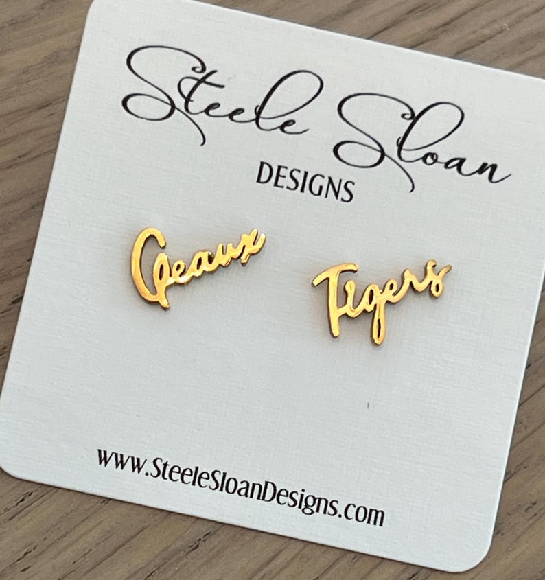May include: Gold-colored "Geaux Tigers" earrings on a white card. The earrings are in a cursive font. The card has the text "Steele Sloan DESIGNS" and the website address www.SteeleSloanDesigns.com.
