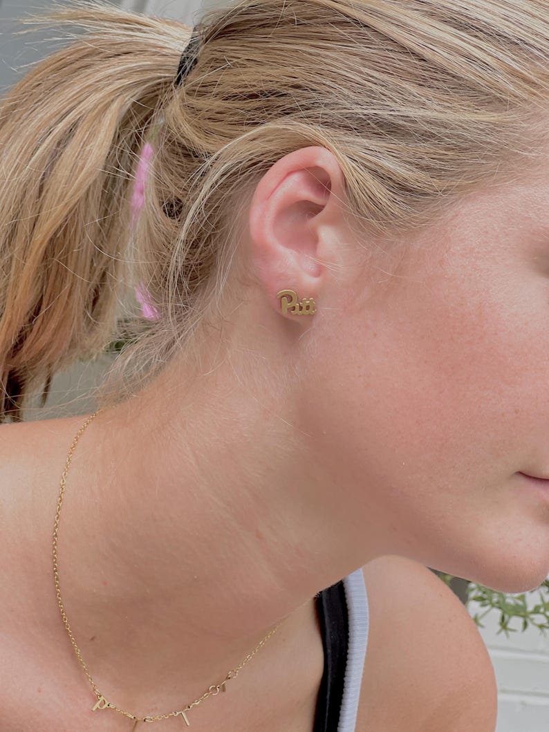 May include: Gold-colored stud earrings with the word "Pitt" in a cursive font. A delicate gold necklace with small letter pendants is also visible. The image focuses on the ear and neck of a person.