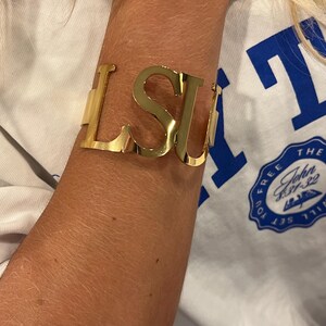 May include: Gold-colored metal cuff bracelet spelling out "LSU" in bold, capital letters. The bracelet is worn on an arm, with a white t-shirt visible in the background. The bracelet is a stylish accessory.