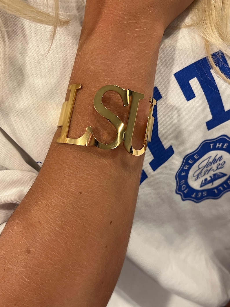 May include: Gold-colored metal cuff bracelet spelling out "LSU" in bold, capital letters. The bracelet is worn on an arm, with a white t-shirt visible in the background. The bracelet is a stylish accessory.