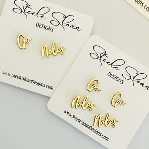 May include: Two white cards with gold lettering that reads "Steele Sloan Designs" and the website address "www.SteeleSloanDesigns.com". The cards display gold earrings with the words "Go" and "Noles" on them.
