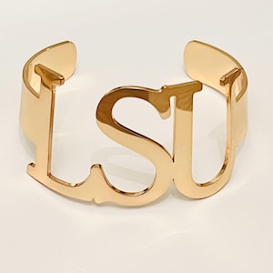 May include: A gold-colored cuff bracelet with the letters "LSU" cut out in a bold, stylized font. The bracelet has a smooth, polished finish and a simple, open design. The letters are connected, forming a single, elegant piece of jewelry.