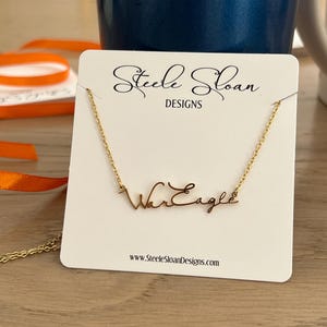Op de afbeelding: Een goudkleurige ketting met de woorden "War Eagle" in een cursief lettertype wordt getoond op een witte kaart. De kaart bevat ook de tekst "Steele Sloan DESIGNS" en een websiteadres. De ketting zit aan een gouden ketting.