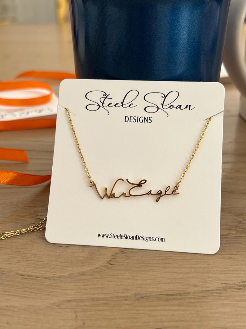 Op de afbeelding: Een goudkleurige ketting met de woorden "War Eagle" in een cursief lettertype wordt getoond op een witte kaart. De kaart bevat ook de tekst "Steele Sloan DESIGNS" en een websiteadres. De ketting zit aan een gouden ketting.
