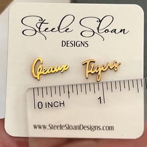 May include: Gold-colored "Geaux Tigers" earrings. The earrings are approximately 1 inch in length. The earrings are displayed on a white card with the text "Steele Sloan Designs" and the website address.