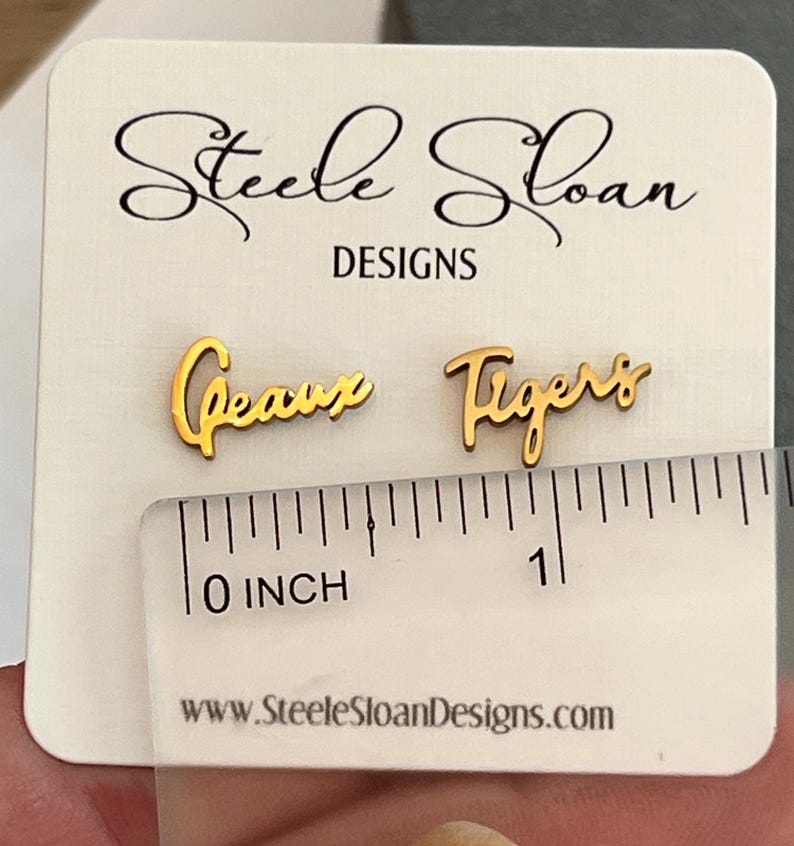 May include: Gold-colored "Geaux Tigers" earrings. The earrings are approximately 1 inch in length. The earrings are displayed on a white card with the text "Steele Sloan Designs" and the website address.
