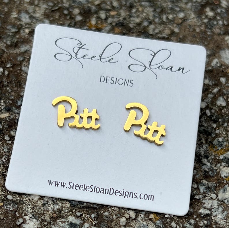 May include: Gold-colored "Pitt" stud earrings on a white card with the text "Steele Sloan Designs" and the website address www.SteeleSloanDesigns.com. The earrings are in a script font. The background is a textured, neutral surface.