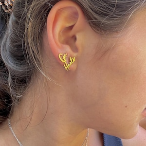 May include: Gold-colored stud earrings spelling out "Go Heels" in a cursive font. The earrings are worn on an ear, with the text clearly visible. The earrings are a stylish accessory.