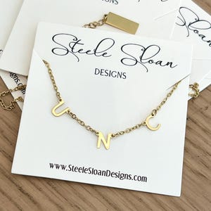 May include: Gold necklace with the letters U, N, and C hanging from it. The necklace is on a white card with the text "Steele Sloan Designs" and "www.SteeleSloanDesigns.com".