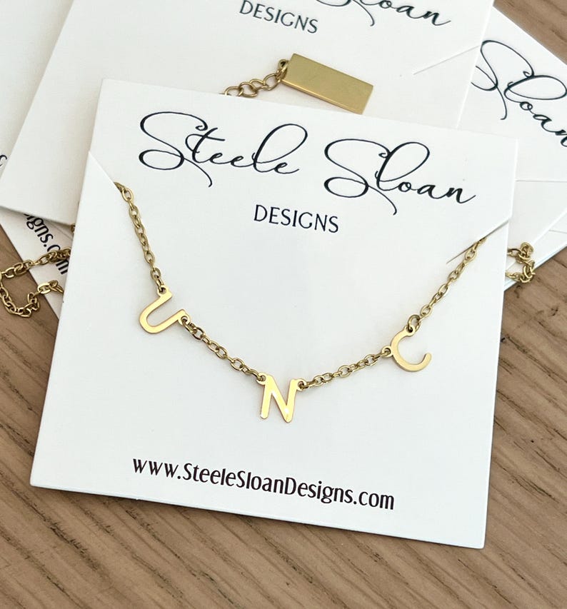 May include: Gold necklace with the letters U, N, and C hanging from it. The necklace is on a white card with the text "Steele Sloan Designs" and "www.SteeleSloanDesigns.com".