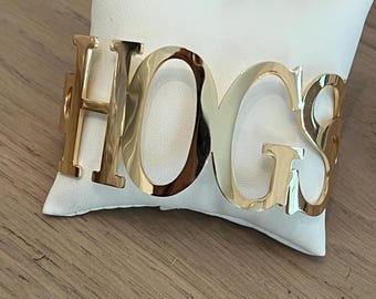 May include: Gold-colored metal bracelet with the word "HOGS" spelled out in large, bold letters.
