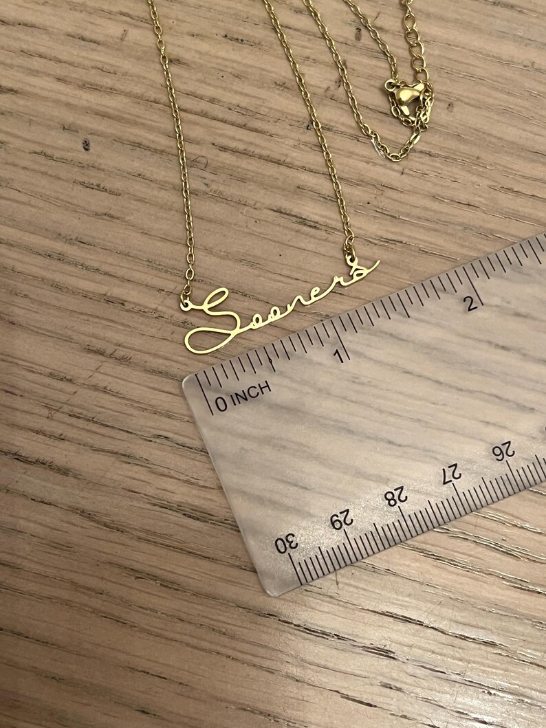 May include: Gold necklace with a cursive script pendant that reads "Sooners". The necklace is measured against a ruler, showing the pendant is about 1 inch long.