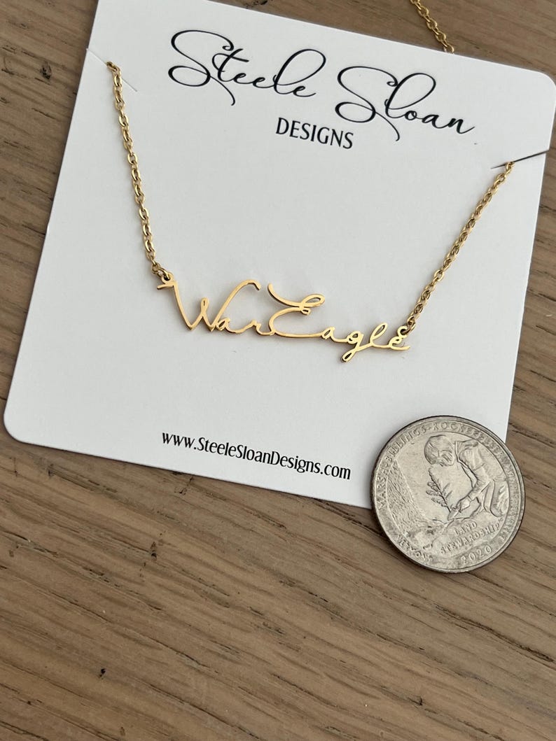 Op de afbeelding: Een goudkleurige ketting met de tekst "War Eagle" in script wordt getoond op een witte kaart. De kaart heeft bovenaan de tekst "Steele Sloan DESIGNS" en onderaan het websiteadres. Een munt ligt naast de kaart.