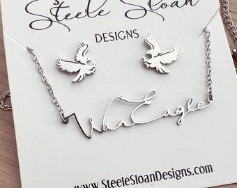 May include: A silver-toned jewelry set featuring a necklace and matching earrings. The necklace spells "War Eagle" in a cursive font. The earrings are shaped like eagles. The set is displayed on a white card with the text "Steele Sloan Designs" and the website address.