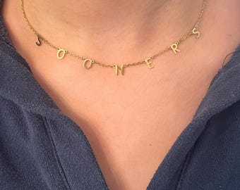 May include: A gold chain necklace with the word "SOONERS" spelled out in individual letters.