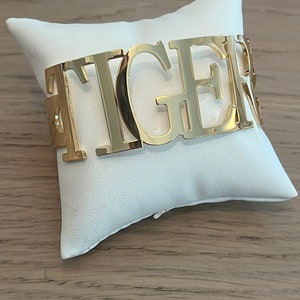 May include: Gold-toned metal bracelet with the word "TIGER" spelled out in large, bold letters.